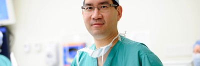 Doctor in green surgical scrubs wearing glasses and cap looking at camera.